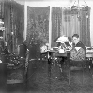 White woman sitting at table in room with hanging light fixture and desk with typewriter