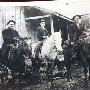 Three white men on horseback outside single-story building with covered porch