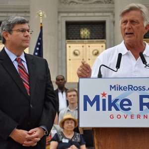 Old white man in white shirt speaking at lectern with white man in suit and tie standing next to him at Capitol building