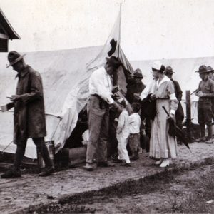 Group of white soldiers in uniform and nurses at camp with row of white tents and children