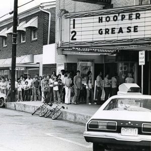 Mixed crowd of people in line outside multistory brick building and theater with "Hooper" and "Grease" on its marquee