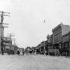 Street lined with buildings and telephone poles with crowd of people and wagons