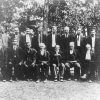 Group of old white men in suits sitting and standing with trees in the background