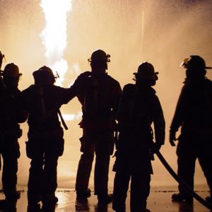 Six firefighters stand spraying hose silhouetted in front of fire at night