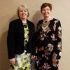 Older white woman smiling in black coat standing next to older white woman with glasses in floral dress