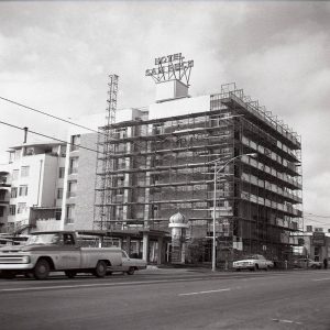 Traffic passing by multistory hotel building with neon sign on top and scaffolding around it on city street