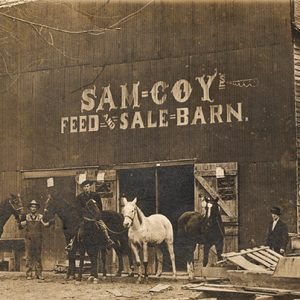 White men with horses and dog standing outside "Sam Coy Feed and Sale Barn" building