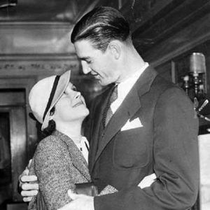 White man in suit and slicked-back hair and tie looking down at and hugging a white woman in hat who is looking up at him