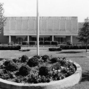 Multistory modern building with short columns high windowless face lawn three flagpoles and round garden