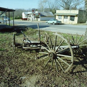 Remnants of wooden wagon and wagon wheel buried in the ground next to storefront on rural town road