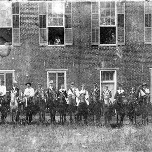 White men on horseback in front of multistory brick building