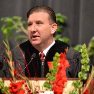 White man speaking at lectern in graduation robes