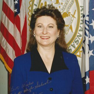 White woman with long hair smiling in blue top with flag and seal behind her