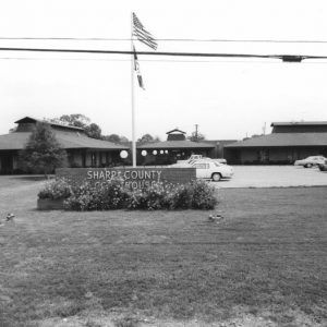 Single-story building with parking lot and flagpoles behind brick sign reading "Sharp County Courthouse"