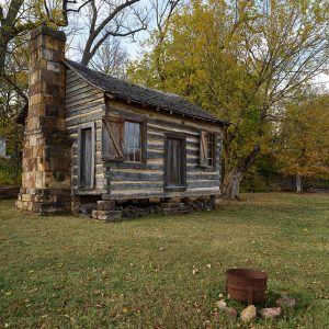 Log cabin with stone block chimney on brick foundations with trees behind it