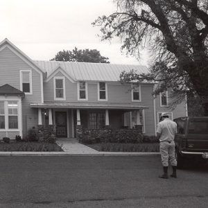 Two-story house with covered porch and man with truck in street under a large tree