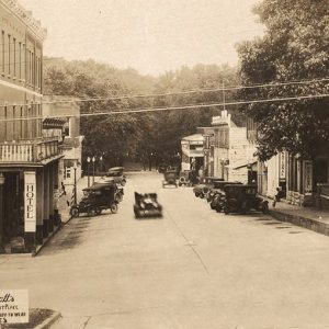 Cars parked on street outside multistory hotel building across from brick storefronts with trees in the background