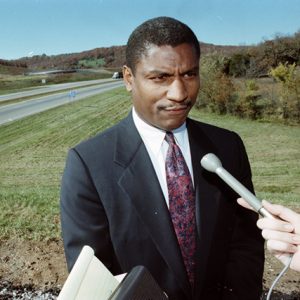 African-American man in suit and tie speaking into a microphone near interstate