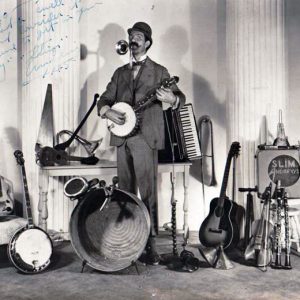 White man with mustache playing a horn and banjo surrounded by many musical instruments