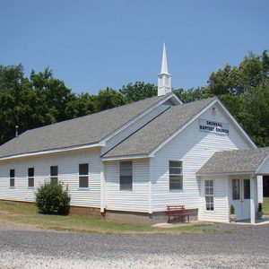 Single-story church building with steeple with gravel in front with sign saying "Snowball Baptist Church"