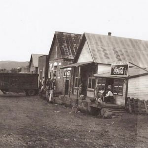 Car and semi-truck parked outside row of single-story storefronts on dirt road