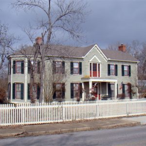 large two-story house with green siding and white fence with red doors and trees in the yard on street