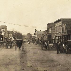 Cars and horse drawn carriages on street with multistory buildings on both sides