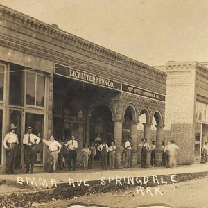 Group of white men and boys standing on sidewalk outside "Lichlyter News Company" building on town street