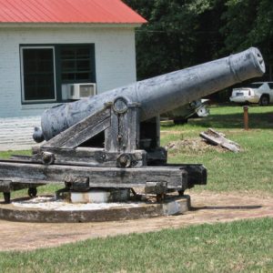 Cannon on round pedestal on concrete platform with single-story white building behind it