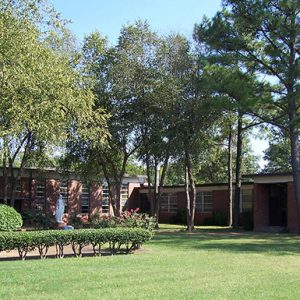 Brick building with rectangular windows and grounds that include bushes and trees