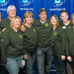 Group of white men and women in matching green shirts and one in white shirt posing with blue background