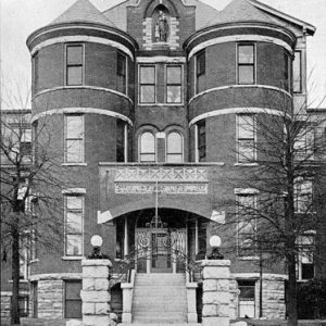 Multistory brick building with circular towers and arched entrance with staircase and brick wall in the foreground