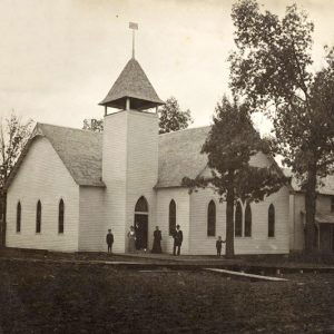 Single-story building with bell tower and gothic arch windows next to multistory house