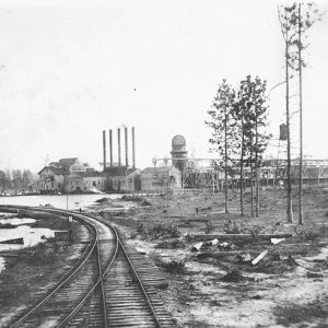 Multistory industrial buildings with four smokestacks and railroad tracks in the foreground