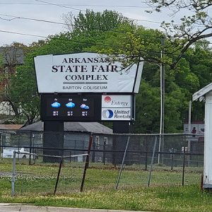 Multistory houses behind electronic Arkansas State Fair Complex sign with fence with single-story outbuilding in the foreground