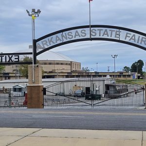 "Arkansas State Fair" arch sign over double gate with fence with single-story building and enclosed stadium in the background