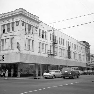 Cars driving on city street by multistory department store with people walking on the sidewalk under awning