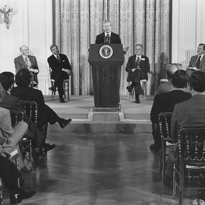 White man speaking at lectern to crowd of white men in suits