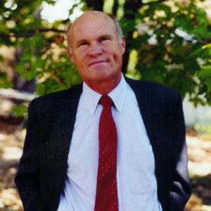 Bald white man in blue suit and red tie with trees in the background