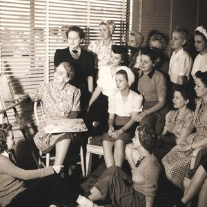 Old white woman sitting at easel teaching group of young white women how to paint in classroom