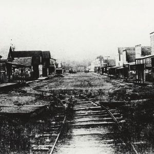 Rows of storefront buildings with covered entrance across from multistory buildings on dirt road with trolley tracks