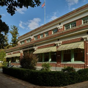 Two-story red brick building with awnings over the first-floor windows and bushes in front