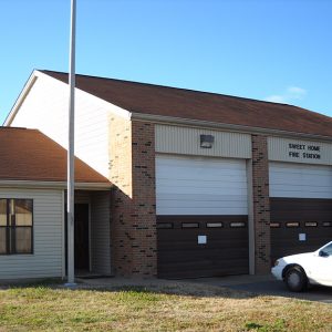 Cars parked at brick fire department building with three garage bay doors