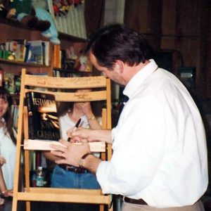 White man in bookshop autographs chair with book "the testament John Grisham" women watching