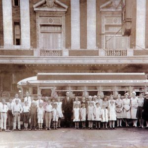 Group of white men women and children posing for a group photo in front of bus outside multistory theater building