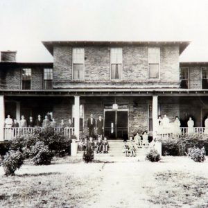 Group of people standing on covered porch of multistory brick house with steps on grass