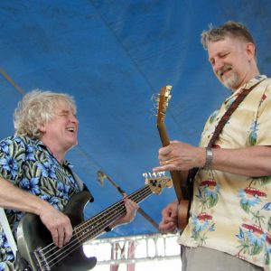 White man singing while playing bass guitar on stage with white man smiling playing electric guitar both under a blue canopy