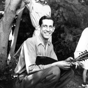 Young white man sitting with guitar amid group of other white men outdoors