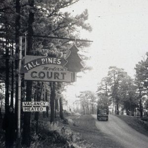 White woman in black standing next to "Tall Pines Motor Inn" sign with truck on road in the background