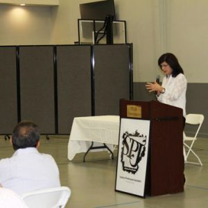 White woman speaking at lectern to indoors crowd